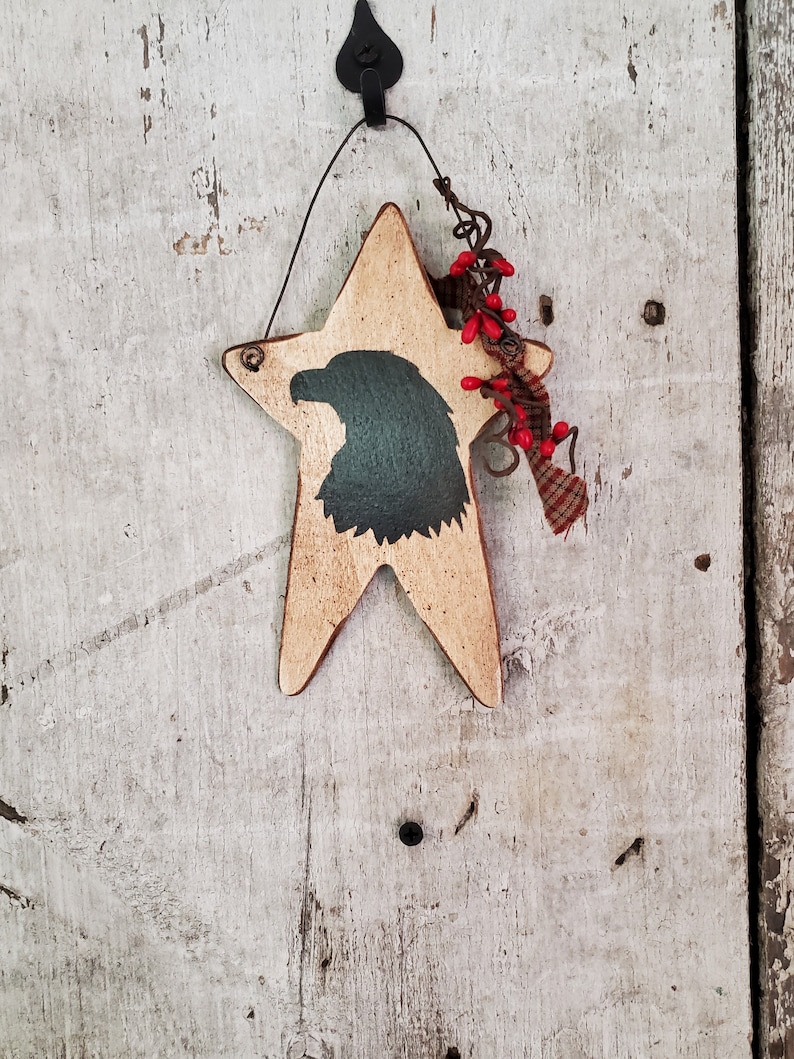 Puede incluir: Un adorno de madera en forma de estrella con una silueta pintada de la cabeza de un &aacute;guila. La estrella est&aacute; pintada de color marr&oacute;n claro y tiene un acabado desgastado. El &aacute;guila est&aacute; pintada de negro y tiene un tono verde. El adorno est&aacute; colgado de un lazo de alambre y tiene una peque&ntilde;a rama de bayas rojas unida a &eacute;l.