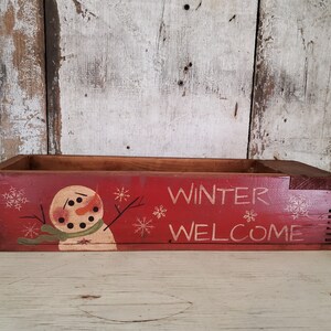 May include: A red wooden box with a painted snowman and the words "Winter Welcome". The box is decorated with snowflakes.