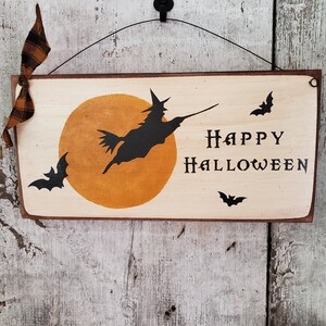 Może przedstawiać: Drewniany znak z namalowanym pomarańczowym księżycem i czarną sylwetką czarownicy lecącej na miotle. Znak głosi "Happy Halloween" z czarnymi nietoperzami wok&oacute;ł tekstu.