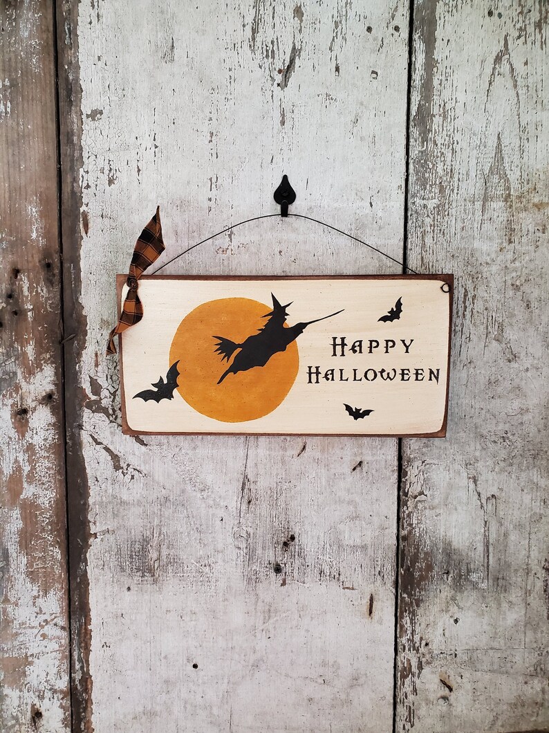 Może przedstawiać: Drewniany znak z czarną sylwetką czarownicy latającej na miotle przed ż&oacute;łtym księżycem. Znak głosi "Happy Halloween" i ma czarne nietoperze wok&oacute;ł krawędzi.