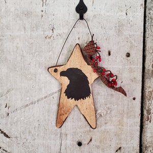 Puede incluir: Un adorno de madera en forma de estrella con una silueta negra de la cabeza de un &aacute;guila. La estrella est&aacute; colgada de un gancho negro y tiene un lazo de tela a cuadros rojo y blanco con bayas rojas.