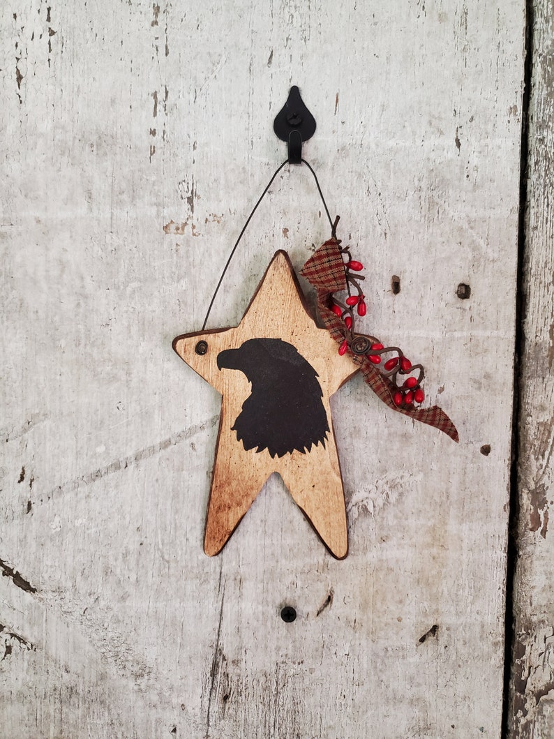 Puede incluir: Un adorno de madera en forma de estrella con una silueta negra de la cabeza de un &aacute;guila. La estrella est&aacute; colgada de un gancho negro y tiene un lazo de tela a cuadros rojo y blanco con bayas rojas.