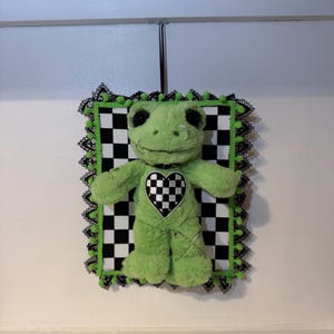 May include: A plush green frog with black eyes and a nose ring, centered on a black and white checkered background. The frog has a checkered heart and is framed with black lace and green pom-poms. Safety pins are used as decoration.
