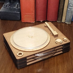 May include: A wooden record player with a light wood top and dark wood sides. The record player is sitting on a wooden table with a glass of amber liquid and a stack of books in the background.