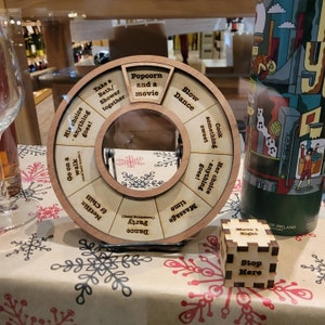 May include: A wooden spinning wheel with a variety of activities to choose from, such as "Take a Bath/Shower Together", "Popcorn and a Movie", "Slow Dance", "Cook Something Sweet", "Massage Time", "Go on a Walk", "Netflix & Chill", and "Party Dance". A wooden cube with the text "Move 1 Right" and "Stop Here" is also visible.