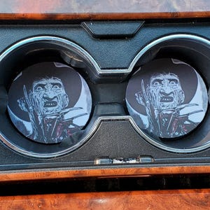 Puede incluir: Dos posavasos para coche con una imagen en blanco y negro de Freddy Krueger. Los posavasos están en los portavasos de un coche, sobre un salpicadero con vetas de madera.