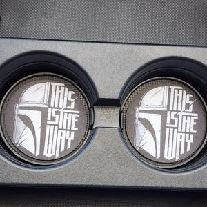 May include: Two black and white car coasters with a Mandalorian helmet design and the text "THIS IS THE WAY". The coasters are round and fit into a car's cup holders. The coasters are made of a durable material.