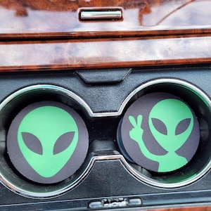 May include: Two black car coasters with green alien designs. One coaster features a full alien face, and the other shows an alien with a peace sign hand gesture. The coasters are in a car's cup holders.