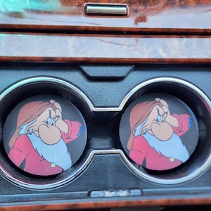May include: Two black car coasters featuring a cartoon image of Grumpy from Snow White. The coasters have a red and white color scheme, with a brown background. The coasters are placed in a car's cup holders.