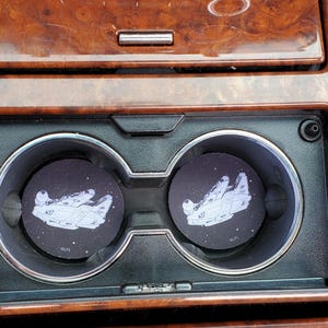 Op de afbeelding: Twee zwarte auto-onderzetters met een witte illustratie van het ruimteschip Millennium Falcon tegen een sterrenachtergrond. De onderzetters liggen in de bekerhouders van een auto, tegen een dashboard met houtnerf.
