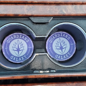 May include: Two round car coasters featuring the text "Guardians of the Galaxy" in a circular design with a stylized star and leaf graphic in the center. The coasters are purple and white.