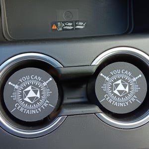 May include: Two black car coasters with white text and graphics. Each coaster has the text "YOU CAN CERTAINLY TRY" around a white geometric shape with two swords. The coasters are in a car's cup holders.