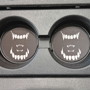 May include: Two black car coasters with a white, stylized monster teeth design. The coasters are round and fit inside a car's cup holders. The cup holders are black.