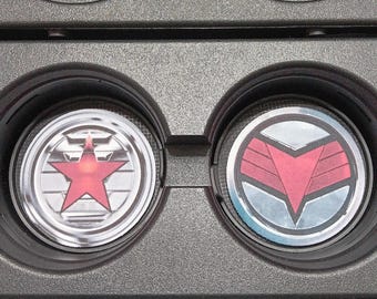 May include: Two car coasters with superhero emblems. One coaster features a red star on a silver background, and the other has a red and silver emblem. The coasters are set in a black car console.