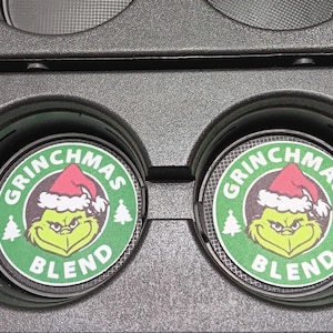 May include: Two car coasters with a Grinchmas Blend design. The coasters are round with a green background and a cartoon Grinch wearing a Santa hat. The text "GRINCHMAS BLEND" is printed around the Grinch image.