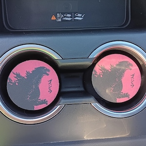 May include: Two pink and black car coasters with a Godzilla silhouette and Japanese text. The coasters are circular and fit in a car's cup holders. The car's interior is visible in the background.