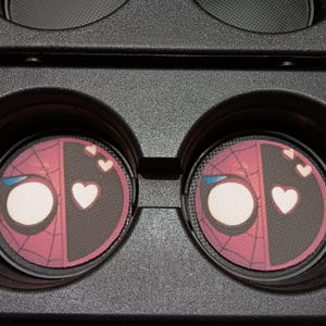May include: Two car coasters featuring a split design: one side depicts Spider-Man's mask, the other Deadpool's mask. Each coaster has a white heart and two smaller hearts. The coasters are set in a black car console.