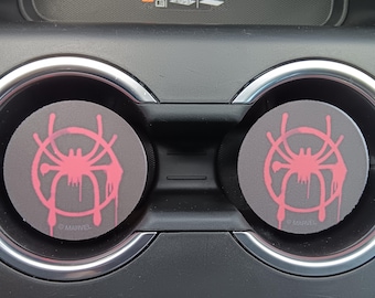 May include: Two gray car coasters with a pink spider graphic and the word "MARVEL" printed on them. The coasters are round and fit into the car's cup holders. The spider graphic has a dripping paint effect.