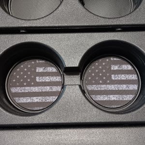 May include: Two black car cup holder coasters with a distressed American flag design. The coasters feature a gray and white stars and stripes pattern, and are set in a black car console.