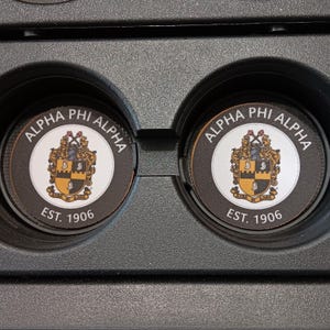 Könnte beinhalten: Zwei runde Untersetzer für das Auto, schwarz und weiß, mit dem Text "ALPHA PHI ALPHA" und einem Wappen. Die Untersetzer sind für die Getränkehalter im Auto konzipiert. Der Text "EST. 1906" ist ebenfalls vorhanden.