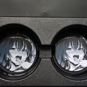 May include: Two car cup holder coasters featuring a manga-style illustration of a woman with black hair and a suggestive expression. The coasters are round and fit into a black cup holder.