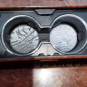 May include: Two silver coins, one with a detailed relief of a woman's face and the other with a detailed relief of a building, rest in a car's cup holders. The coins are set against a wood-grain dashboard.