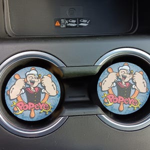 May include: Two round car coasters featuring Popeye the Sailor Man. The coasters have a blue background with a nautical design and the word "Popeye" in pink. Popeye is depicted with his signature sailor hat and flexing his biceps.