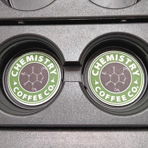 May include: Two coffee pods with the text "CHEMISTRY COFFEE CO." in a green and white circular design. The pods are in a black holder. The design includes a chemical structure.