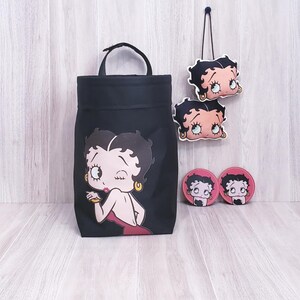 May include: Black tote bag, two coasters, and two hanging ornaments featuring a cartoon woman winking. The tote bag has a handle and a large graphic of the cartoon woman. The coasters and ornaments are pink and black.