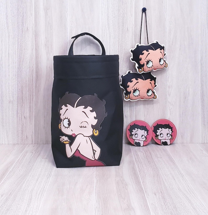 May include: Black tote bag, two coasters, and two hanging ornaments featuring a cartoon woman winking. The tote bag has a handle and a large graphic of the cartoon woman. The coasters and ornaments are pink and black.