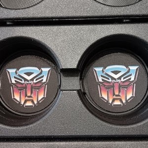 May include: Two black car coasters featuring the Transformers Autobot logo in a gradient of blue, pink, and orange. The coasters are round and fit into a car's cup holders. The logo is centered on each coaster.