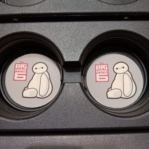 May include: Two gray car coasters featuring the character Baymax from Big Hero 6. Each coaster has a white Baymax illustration and the Big Hero 6 logo in red. The coasters are set in a black car console.