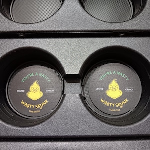 May include: Black car coasters with a yellow Grinch face and the text "YOU'RE A NASTY GRINCH" and "WASTY SKUNK". The coasters are round and fit into a car's cup holders.