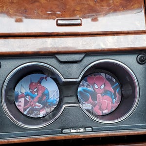 May include: Two round car coasters featuring Spider-Man in action poses. The coasters are placed in the cup holders of a car's center console. The console has a wood grain finish.