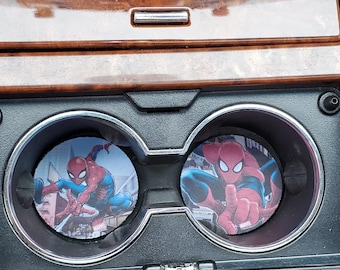 May include: Two round car coasters featuring Spider-Man in action poses. The coasters are placed in the cup holders of a car's center console. The console has a wood grain finish.