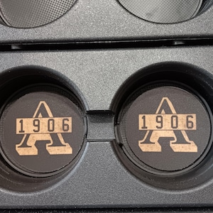 Könnte beinhalten: Zwei schwarze Auto-Untersetzer mit einem beigefarbenen "A"-Logo und der Zahl "1906" aufgedruckt. Die Untersetzer sind kreisförmig und passen in den Getränkehalter eines Autos. Der Hintergrund ist eine schwarze Autokonsole.
