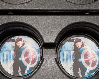 May include: Black VR headset with circular lenses featuring a graphic of a person holding a shield. The shield has a red, white, and blue design. The person is wearing a dark outfit and is set against a blue and white background.