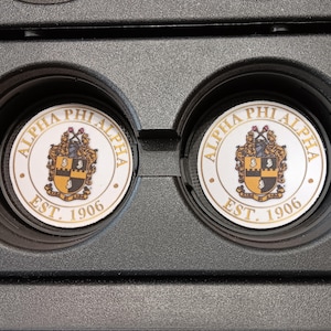 Könnte beinhalten: Zwei weiße und goldene Auto-Untersetzer mit den Worten "ALPHA PHI ALPHA" und "EST. 1906" aufgedruckt. Die Untersetzer haben ein Wappen-Design in der Mitte mit schwarzen, goldenen und weißen Farben. Die Untersetzer befinden sich in einem schwarzen Auto-Getränkehalter.