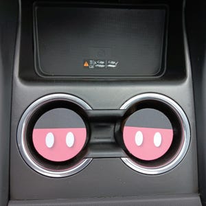May include: Car cup holder coasters featuring a pink and black Mickey Mouse design. The coasters have a white oval shape for the eyes. The coasters are placed in a black car console with a silver trim.