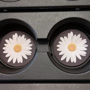 May include: Two car coasters with a daisy design. Each coaster features a white daisy with a yellow center on a dark gray background. The coasters are placed in a black car cup holder.