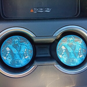 Puede incluir: Dos posavasos azules para coche con una ilustración detallada de un personaje de Mortal Kombat. Los posavasos redondos encajan en los portavasos del coche. La imagen está sobre un interior negro.