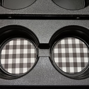 May include: Black car cup holder with two round coasters. The coasters feature a black and white gingham pattern. The cup holder is made of textured black plastic.