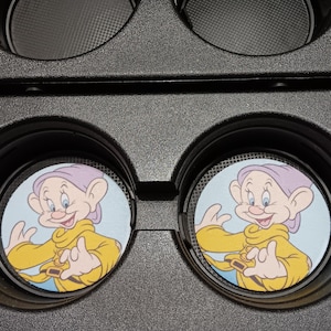 May include: Two car coasters featuring a cartoon character with a purple hat and yellow jacket. The coasters are round with a light blue background and are set inside a black car console.