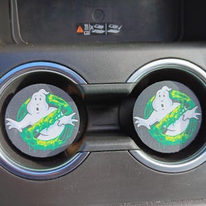 Puede incluir: Dos posavasos para coche con el logo de Cazafantasmas. Cada posavasos tiene un fondo negro con una figura de fantasma blanco dentro de un círculo verde y amarillo. Los posavasos están en los portavasos de un coche.