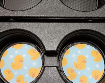 May include: Two car coasters with a light blue background and a pattern of yellow rubber ducks and white bubbles. The coasters are placed inside a black car console cup holder.