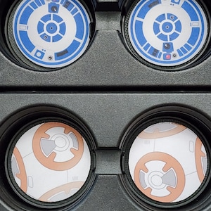 May include: Four round coasters featuring Star Wars droid designs. Two coasters display a blue and white BB-8 design, while the other two showcase an orange and white BB-8 design. The coasters are set in a black holder.