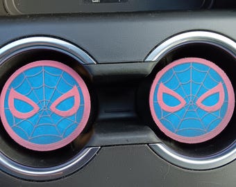May include: Two round car coasters featuring a blue spiderweb design with a pink outline of a mask. The coasters are placed inside the car's cup holders. The coasters are a fun and functional car accessory.