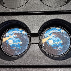 May include: Two car coasters featuring a Godzilla design in the style of Van Gogh's "Starry Night." The coasters have a blue, yellow, and black colour scheme, with a monster breathing fire. The coasters are round and fit in a car's cup holder.