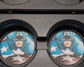 May include: Two round car coasters featuring Captain America. The coasters have a blue, white, and red design with the letter "A" and a star. The coasters are placed in a black car console.