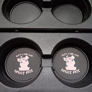以下が含まれることがあります： 豚のグラフィックと「Just a girl who loves pigs」の文字が入った黒い車のコースター。豚は薄いピンク色で、花の冠が付いています。コースターは車のカップホルダーに合うように設計されています。